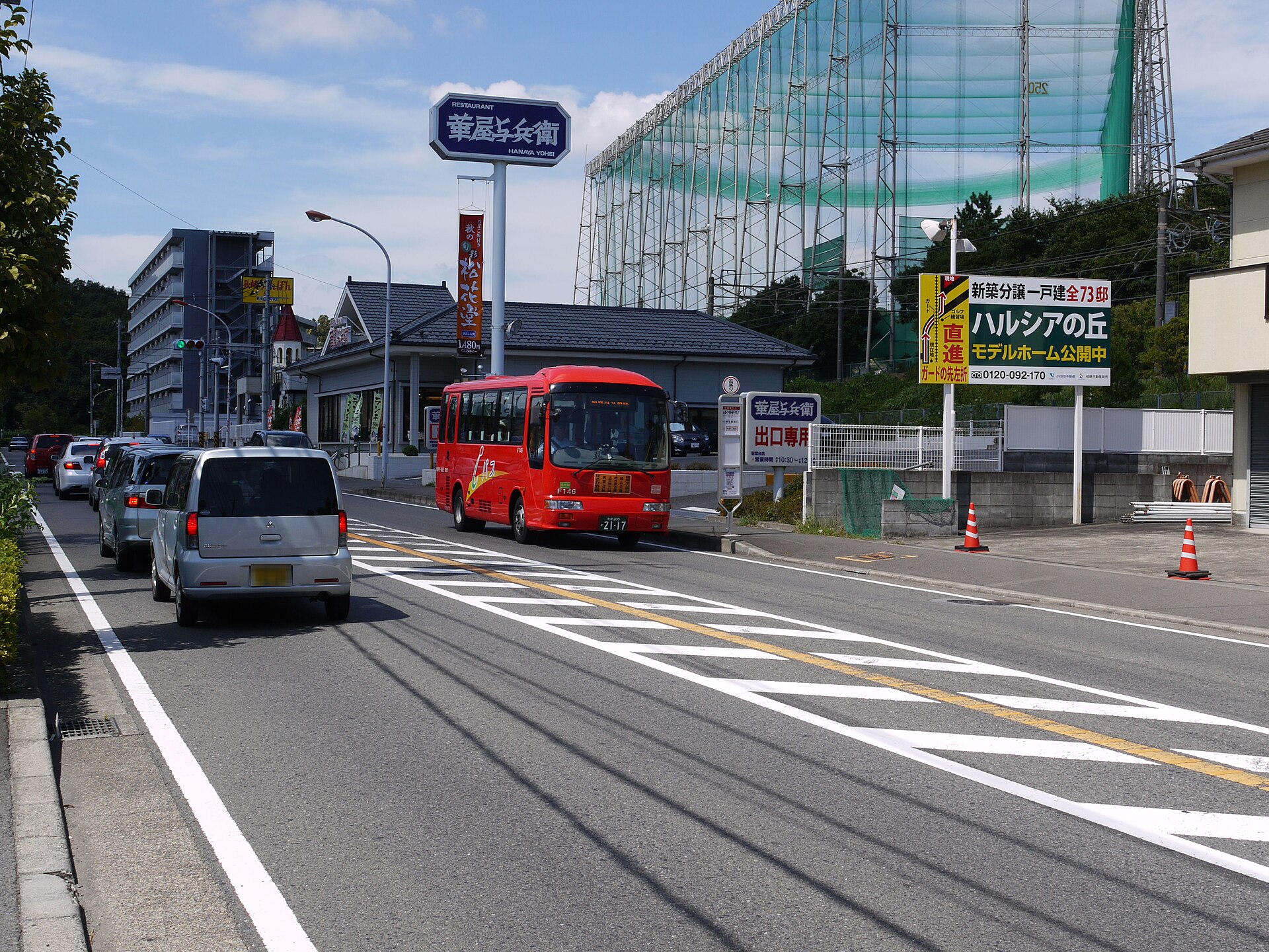 道路わきのバス停の標識の近くを走る、赤い小型のバス。標識の上に「はるひ野駅入口」と読める