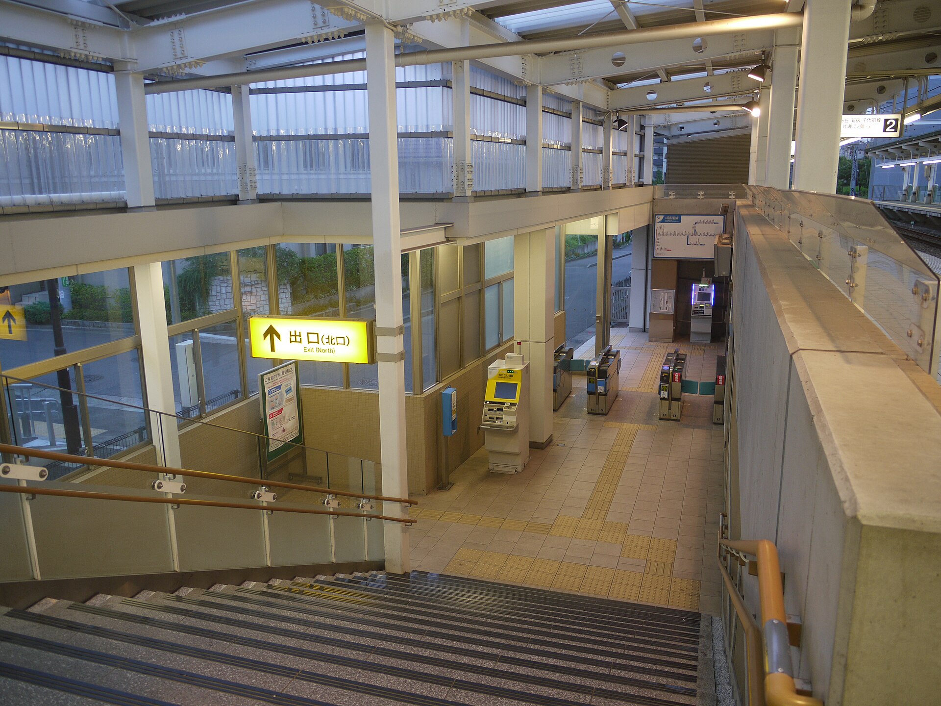 はるひ野駅の北口の改札を、階段の上から見下ろした様子。黄色い「出口（北口）」の看板と、券売機、改札がある