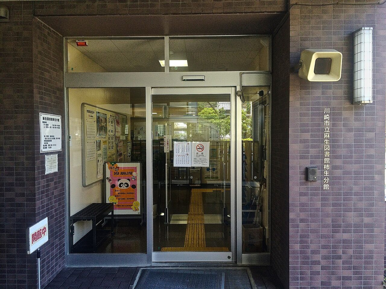 麻生図書館柿生分館の入口。ガラスの自動ドアの左に、開館時間の案内と、おはなし会のポスターが見える